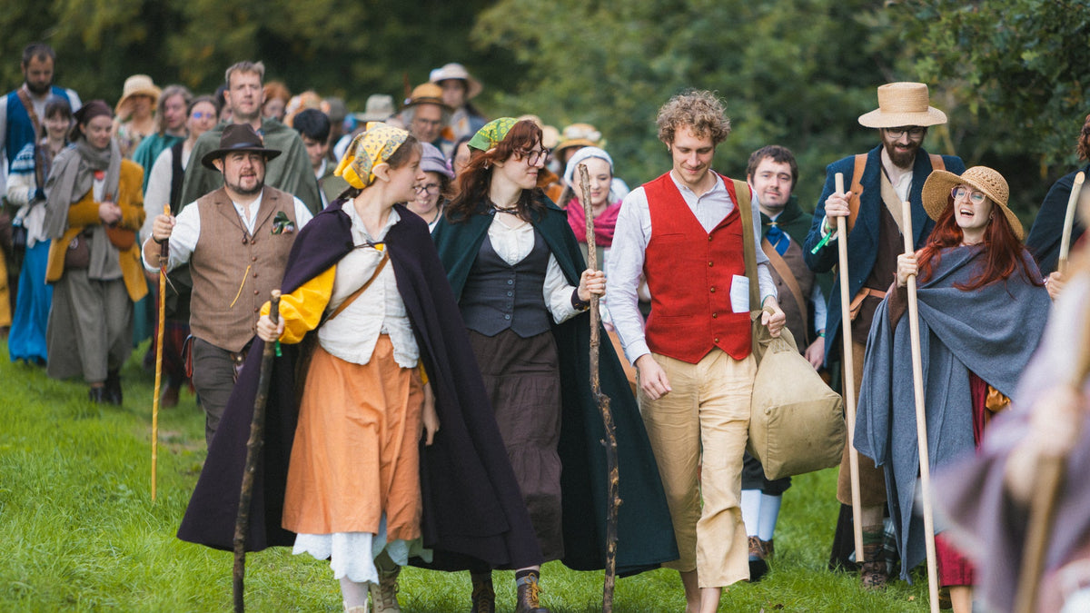 A group of people dressed in medieval or historical costumes walking along a grassy outdoor path, carrying staffs and wearing colorful garments including a red vest, tan skirt, and dark cloaks. Brandywine Hobbit Festival Larp Burgschneider