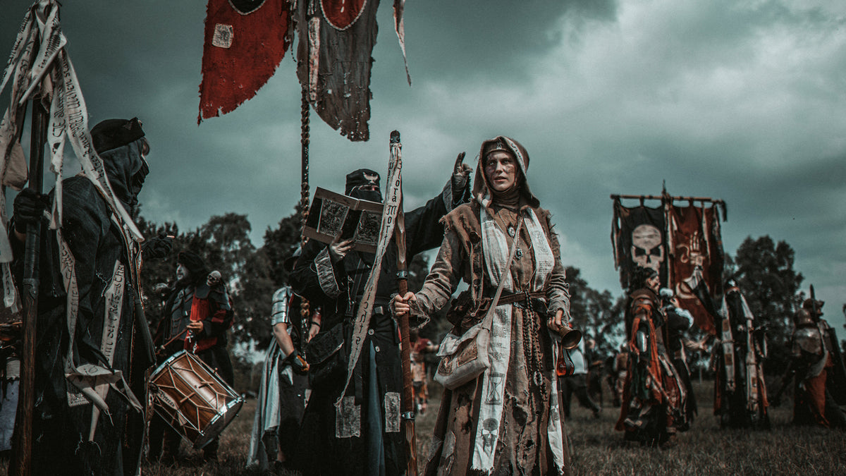 A dramatic outdoor scene showing a group of medieval or Viking-style figures in dark cloaks and armor, carrying banners, flags, and a drum under a stormy gray sky. Mythodea Larp Burgschneider