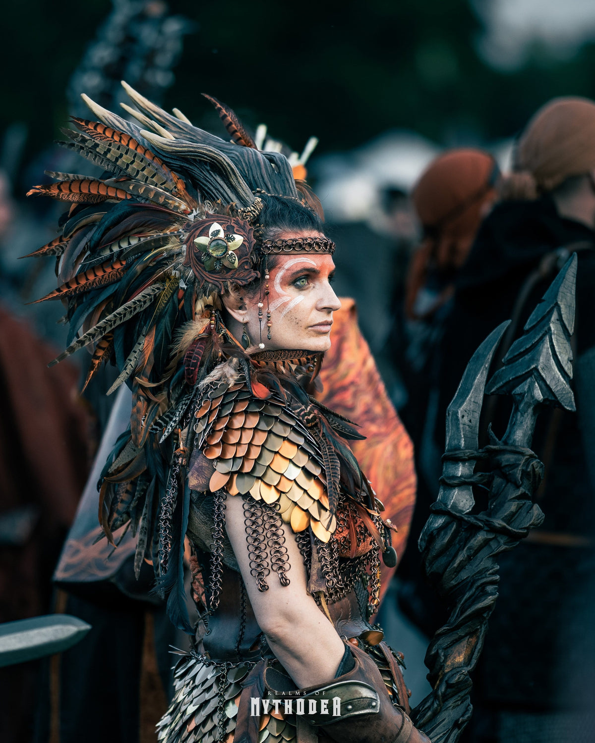 A person in elaborate fantasy costume shown in profile, wearing a large feathered headdress, scale-like armor pieces in dark brown, bronze, and black, with face paint and a blurred outdoor crowd background. Mythodea LARP Burgschneider
