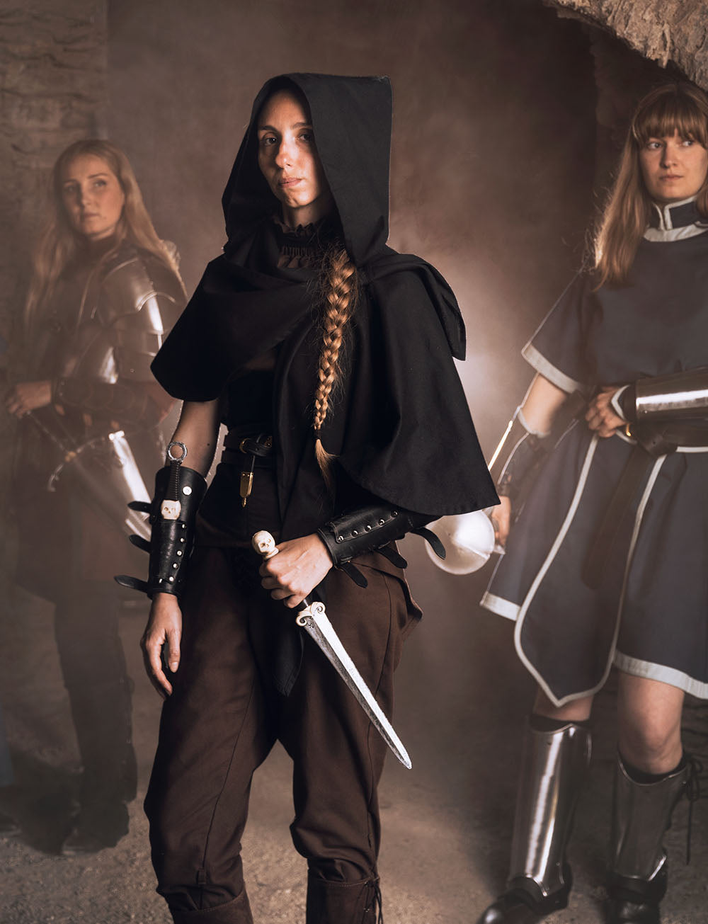 Person in dark D&D medieval attire holding a sword with two other individuals in similar costumes in the background.
